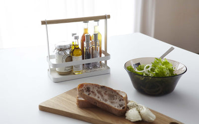 White metal spice rack holding spice canisters and oils on a kitchen countertop next to salad and bread.