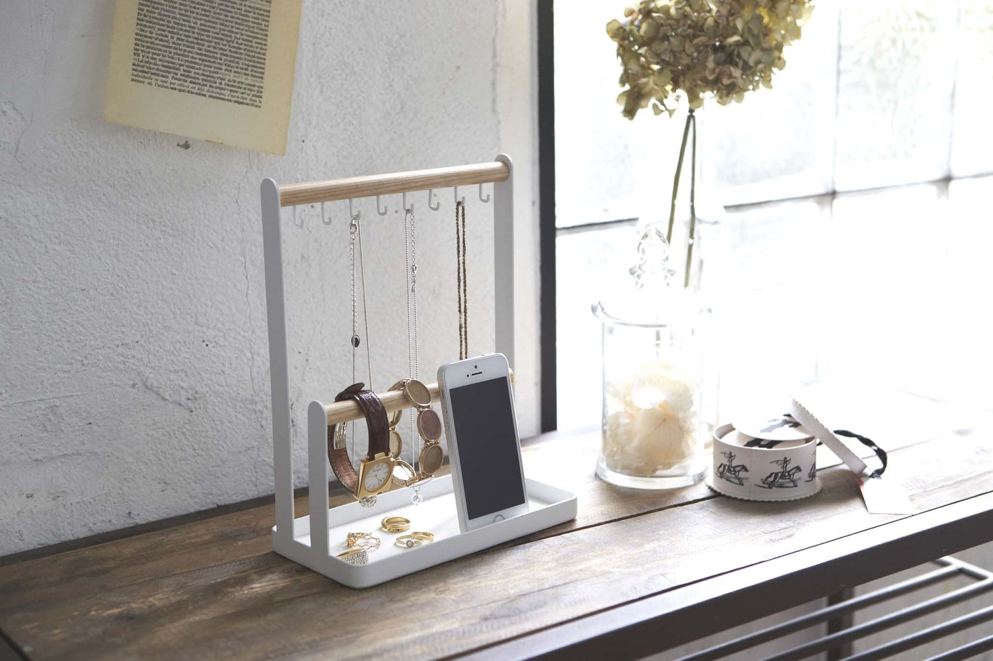 Wood and steel accessories tray filled with necklaces, watches, and mobile phone; sitting on a desk in a sunny room.