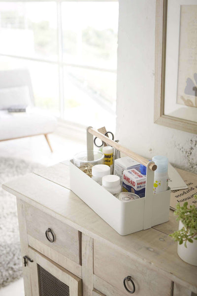 Rectangular white Yamazaki toolbox with wooden handle containing household items, sitting on dresser.